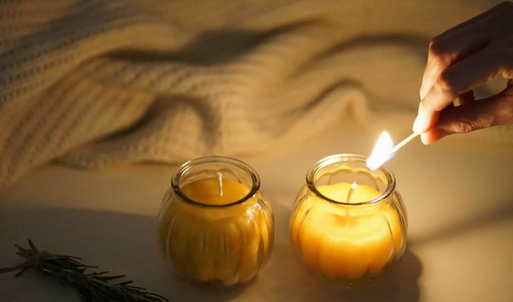 two beeswax candles in glass jars, a cream sweater and a hand holding a burning match