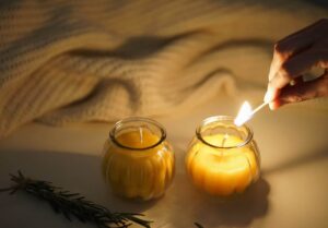 two beeswax candles in glass jars, a cream sweater and a hand holding a burning match