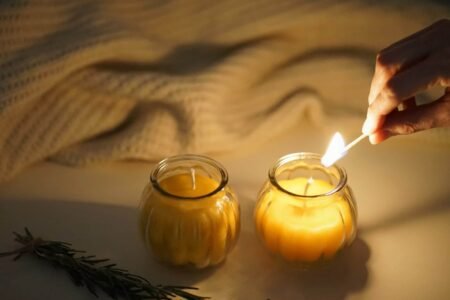 two beeswax candles in glass jars, a cream sweater and a hand holding a burning match
