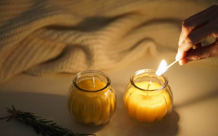two beeswax candles in glass jars, a cream sweater and a hand holding a burning match