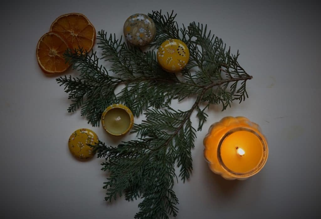 a burning candle in a glass jar, three mini tin candles, a pine branch and two orange slices