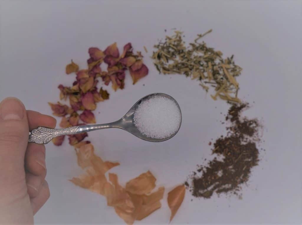 a hand holding a spoon with alum above several natural dye materials (onion skins, madder, fustic, rose petals)