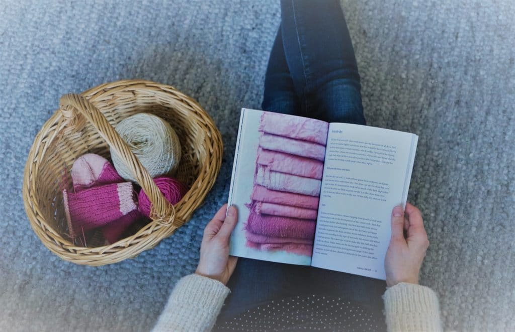two hands holding a book about botanical dyeing next to a wooden basket with wool and a sock knitting project