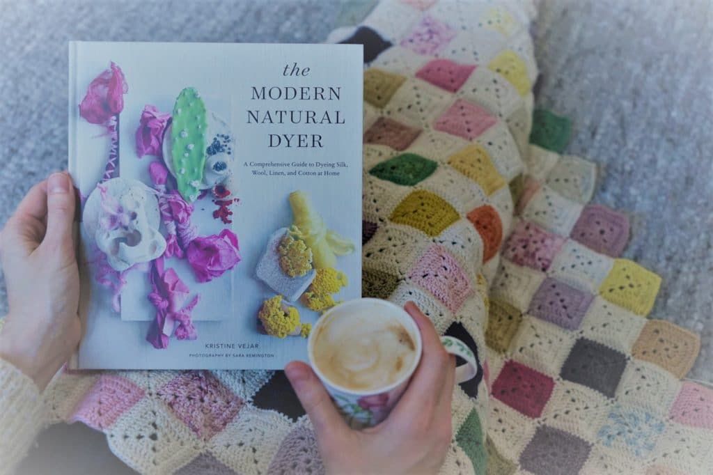 two hands holding the book ther modern natural dyer by kristine vejar and a cup of coffee on top of a crocheted granny square balnket