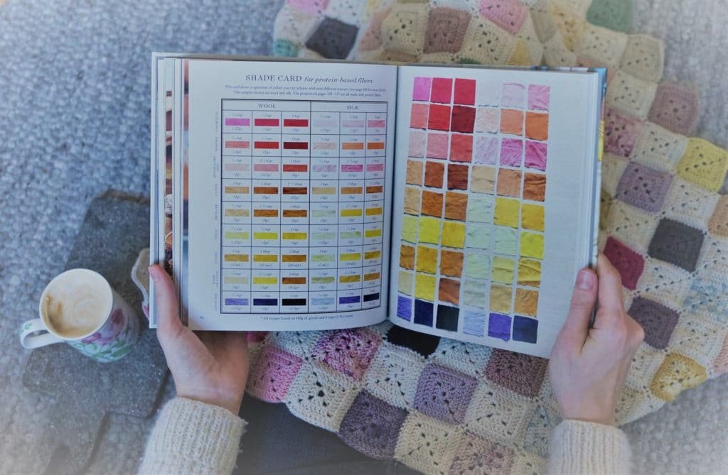 two hands holding a book about natural dyeing on top of a crocheted granny square blanket and a cup of cofee sitting netx to the person