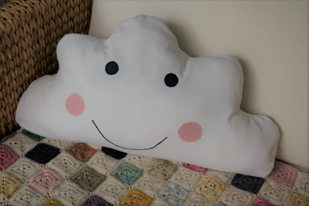cloud pillow with a face embroidered on it sitting in a chair alongside a crochet blanket