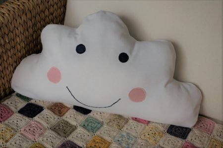 cloud pillow with a face embroidered on it sitting in a chair alongside a crochet blanket
