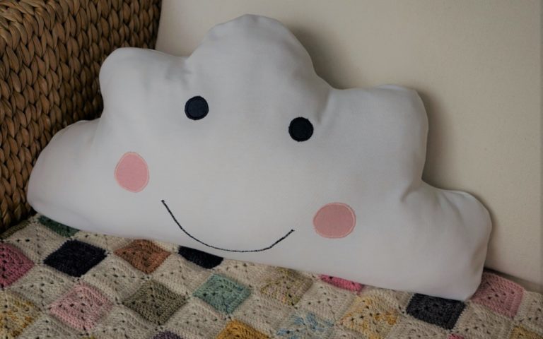 cloud pillow with a face embroidered on it sitting in a chair alongside a crochet blanket