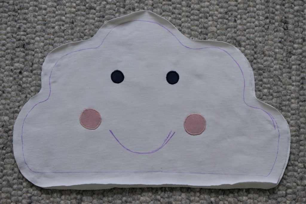 a cloud shape drawn onto a white fabric and four circles in pink and blue for the cheeks and eyes laying on it