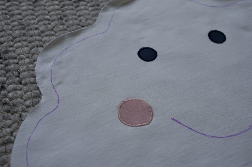 a cloud shape drawn onto a white fabric and four circles in pink and blue for the cheeks and eyes laying on it
