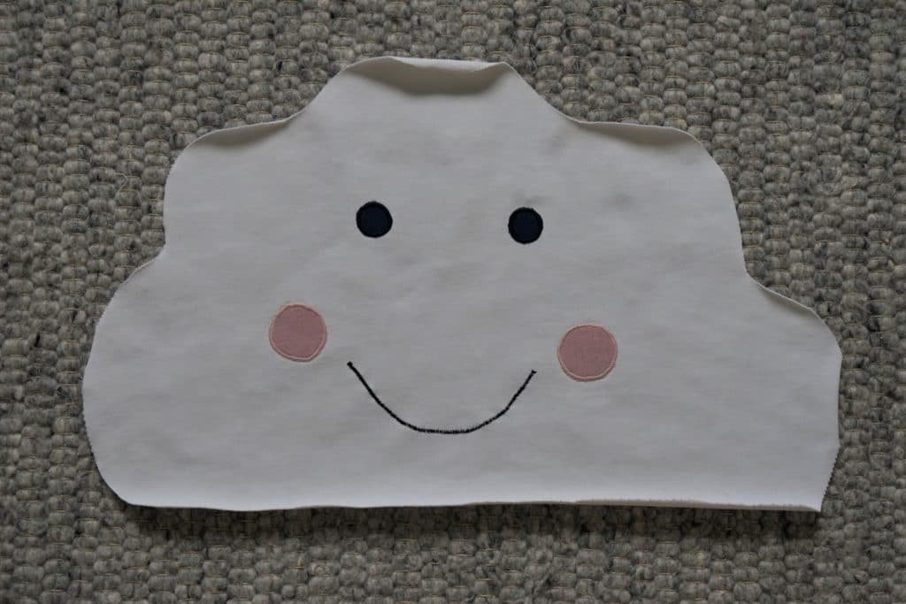 a cloud shape drawn onto a white fabric with eyes, cheeks and a mouth appliquéd on it