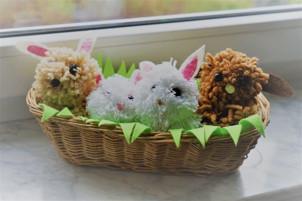 four Easter bunnies made out of pom poms sitting in a wooden basket with some green felt that looks like grass