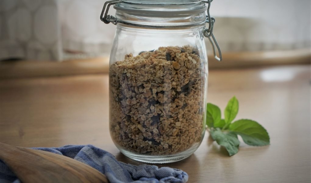 homemade granola in a glass container, a wooden spatula, a towel and some apple tree leaves