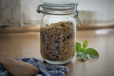 homemade granola in a glass container, a wooden spatula, a towel and some apple tree leaves
