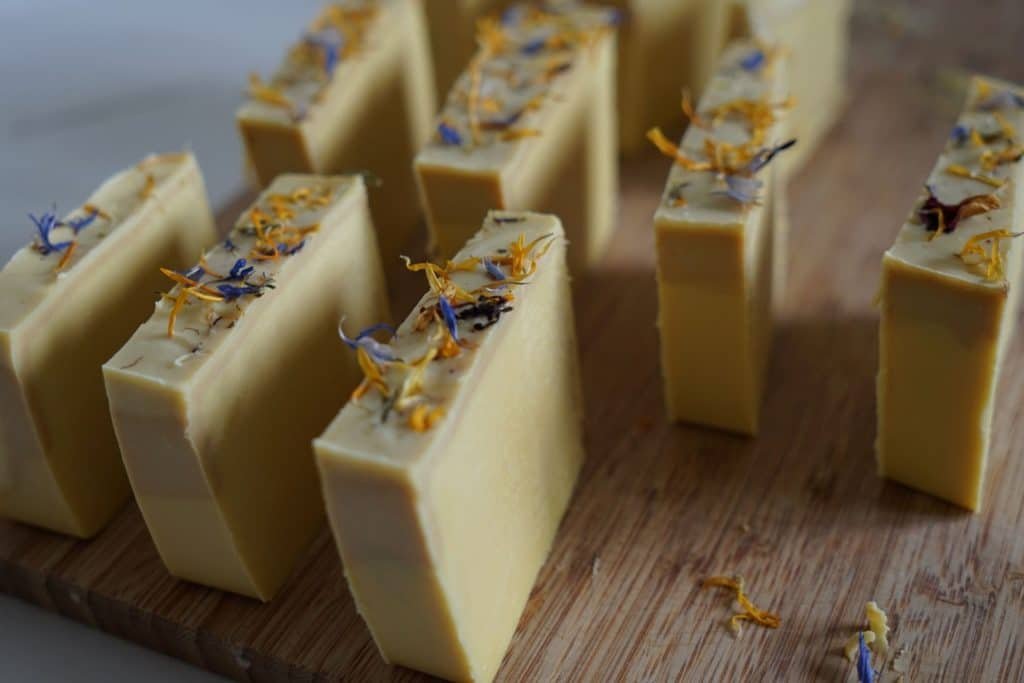 loaf of yellow cold process soap topped with wildflowers cut into pieces on a wooden cutting board