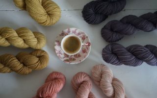 a floral cup of espresso sitting in the middle of several skeins of yarns in shades of pinks, purples and browns