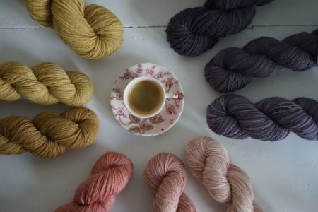 a floral cup of espresso sitting in the middle of several skeins of yarns in shades of pinks, purples and browns