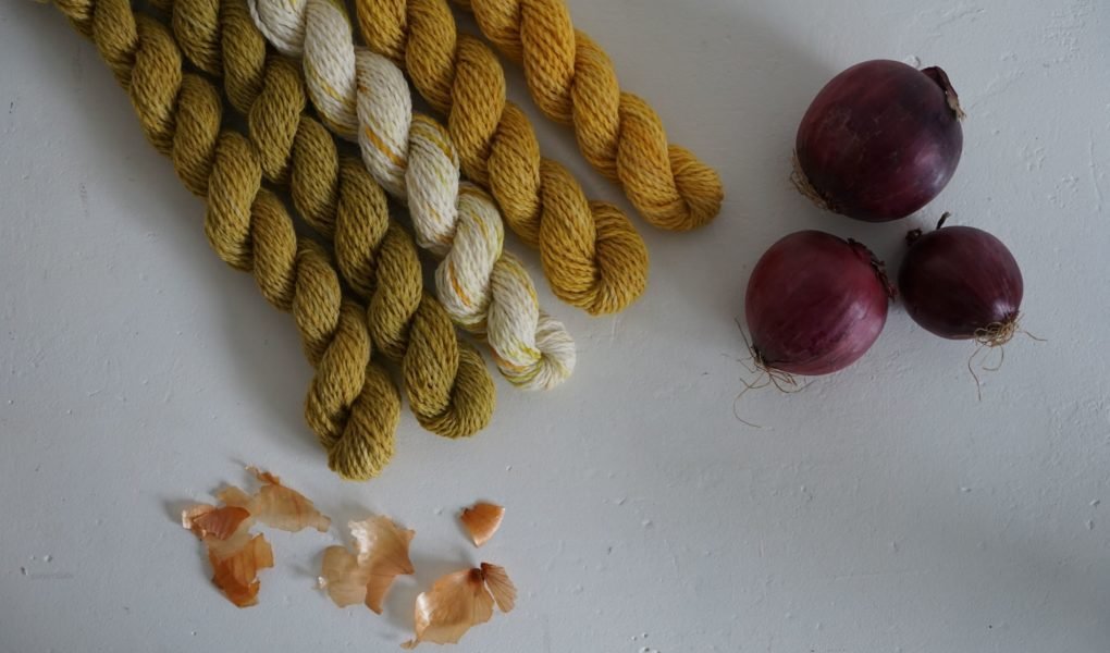 five mini skeins of yarn naturally dyed with onion skins alongside some yellow onion skins and three red onions