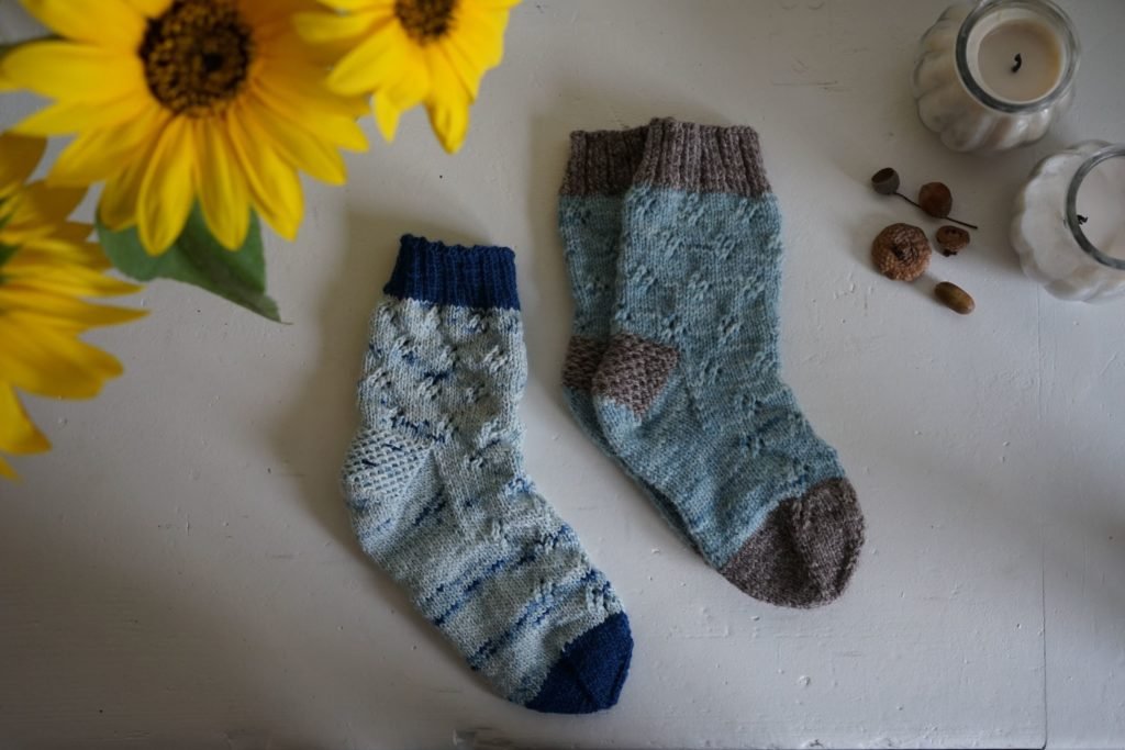 three blue hand knitted wool socks on a white bench next to a sunflower and some candles
