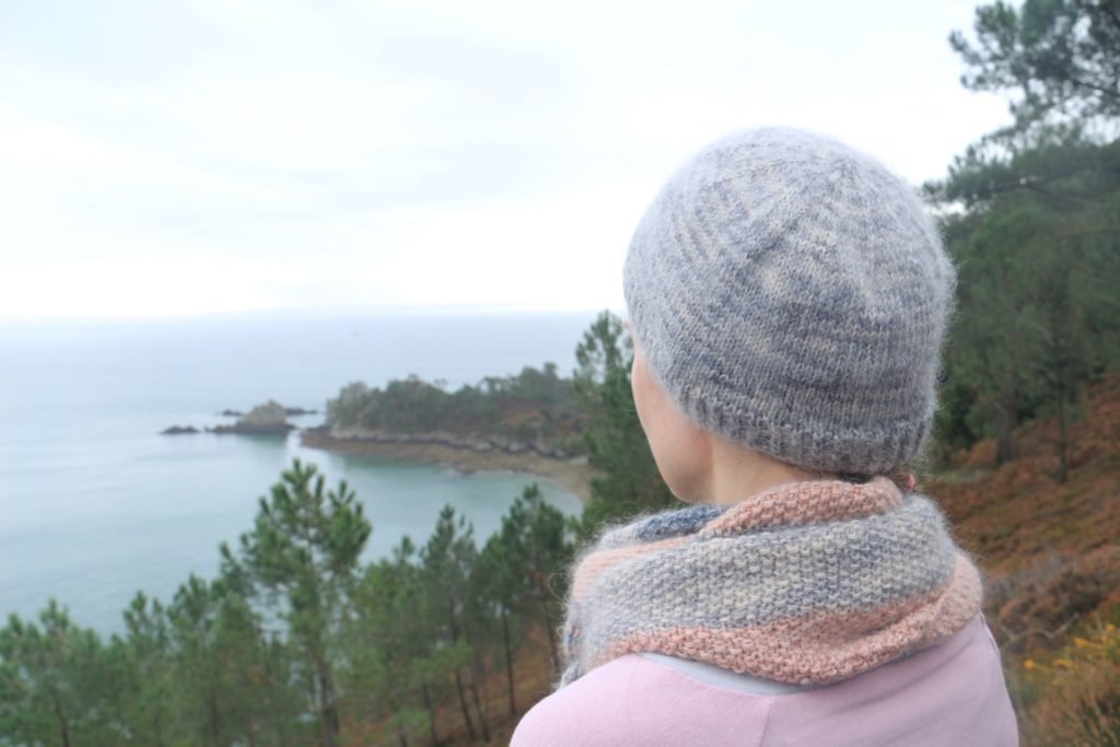 woman standing on the side of a cliff, dressed in knitwear, with a stunning view of the ocean and landscape