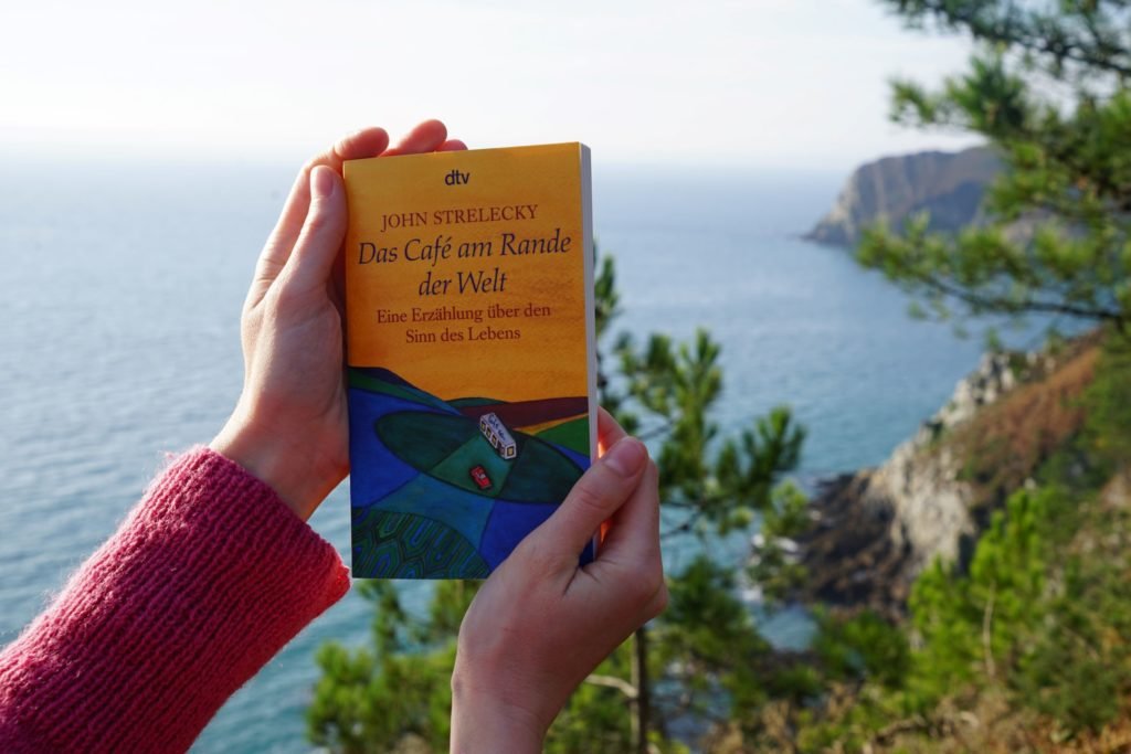 two hands holding a copy of the why café with a stunning ocean view in the background