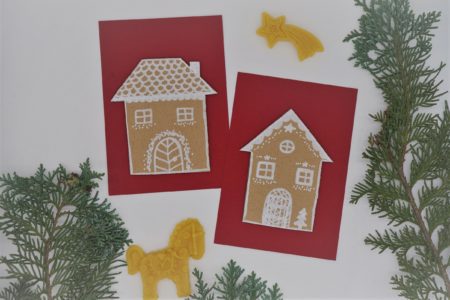 two red card with cardboard gingerbread houses on them next to some beeswax ornaments and branches