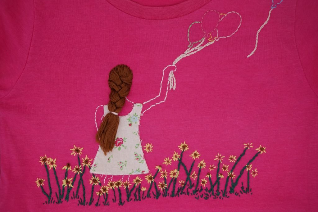 pink t shirt with an embroidery of a girl with brown hair in a field of sunflowers holding some balloons