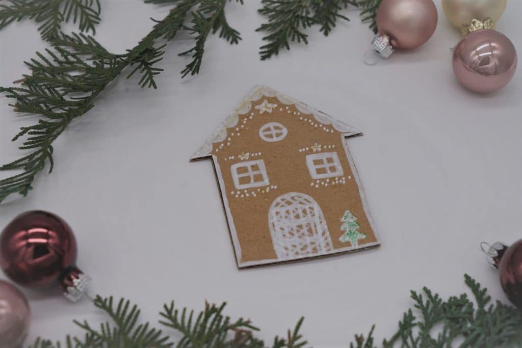 gingerbread house made from cardboard next to some branches and mini Christmas ornaments