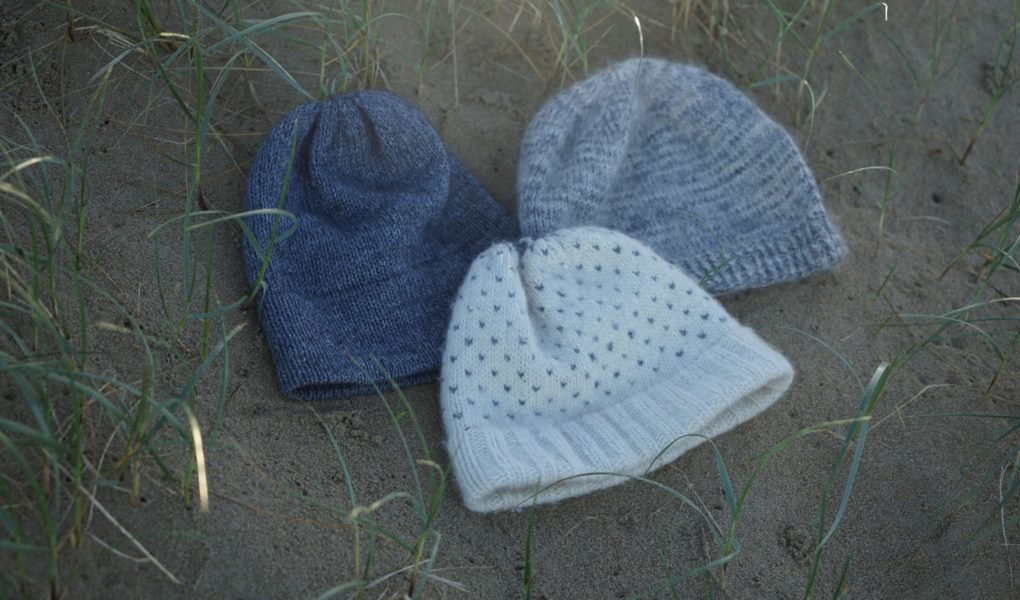 three knitted hats in blue, greys and white laying on a sand dune