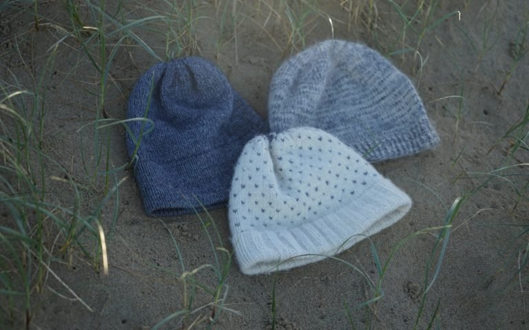 three knitted hats in blue, greys and white laying on a sand dune
