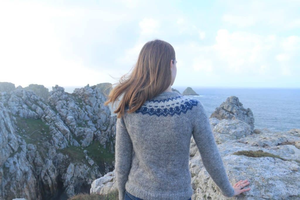 back of a woman with dark blonde hair standing on a cliff at the ocean wearing the strokkur sweater pattern