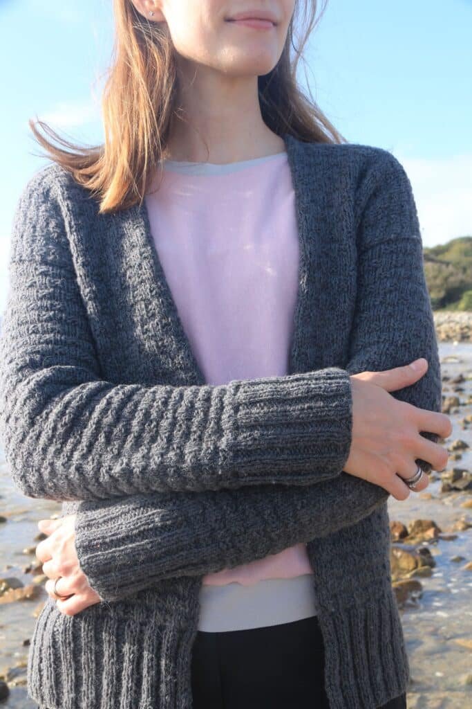 a woma standing on a beach wrapped in a handknitted grey cardigan