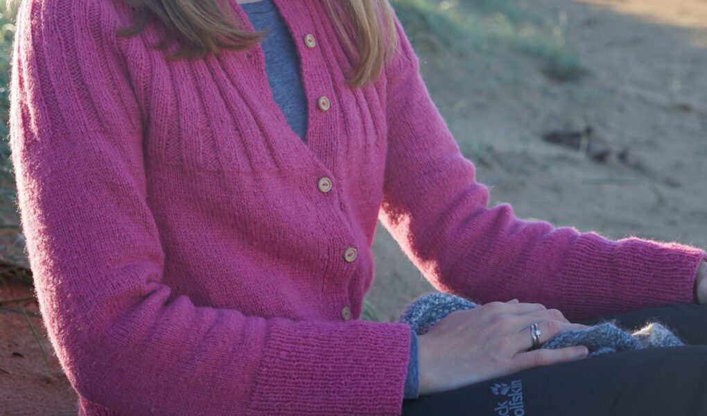 a woman sitting on a beach wearing a pink handknitted cardigan