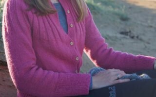 a woman sitting on a beach wearing a pink handknitted cardigan