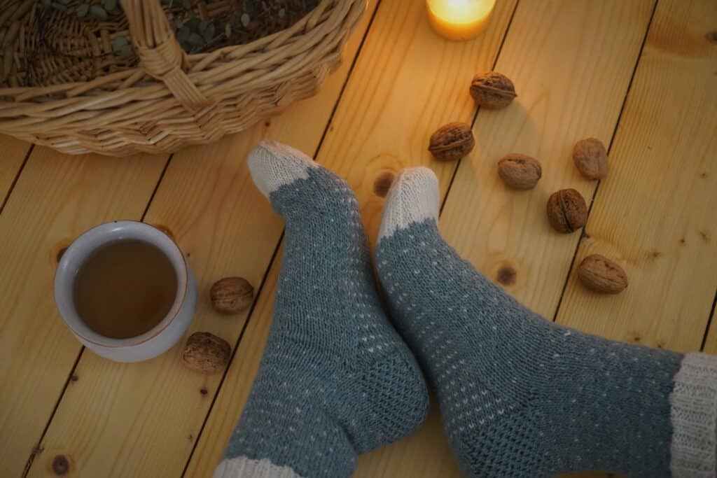 two feet in handknitted blue and white sockes next to each other on a wooden floor next to a cup of tee, some walnuts, a basket and a burning candle