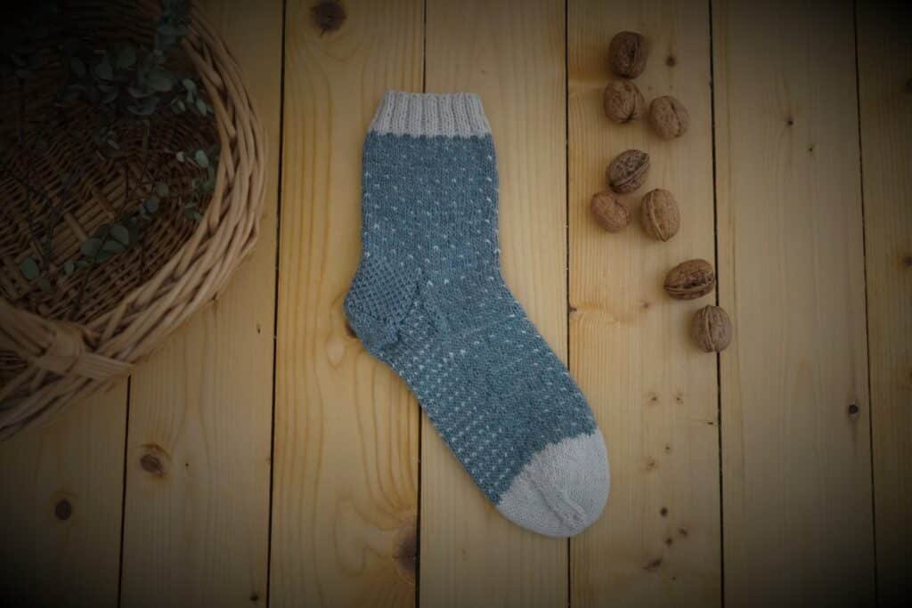 a blue and white handknitted sock on a wooden floor next to a basket and some walnuts