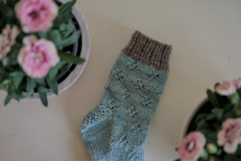 one handknitted blue and grey sock on a white background with some pink flowers next to it