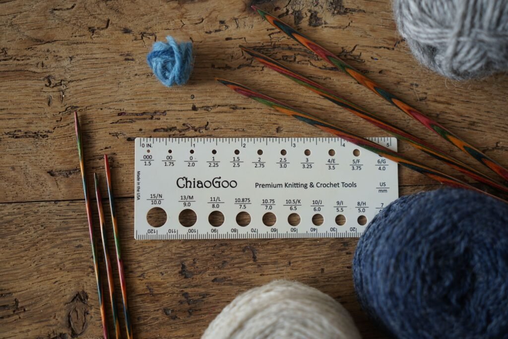 a needle size tool, knitting needles and various balls of yarn laying on a a wooden surface