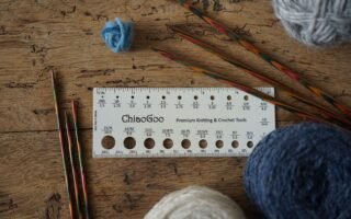 a needle size tool, knitting needles and various balls of yarn laying on a a wooden surface