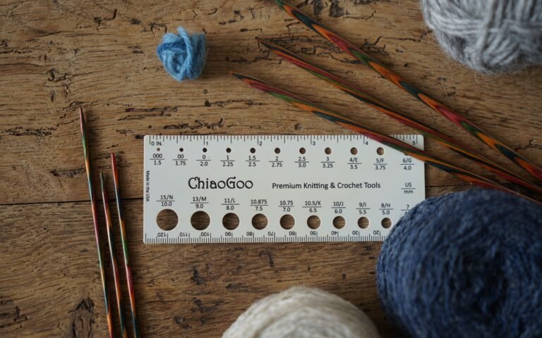 a needle size tool, knitting needles and various balls of yarn laying on a a wooden surface