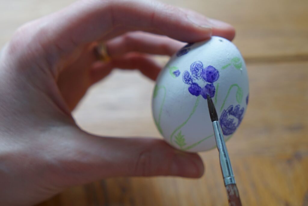 a hand holding a hand painted easter egg and a paintbrush adding water to the painting