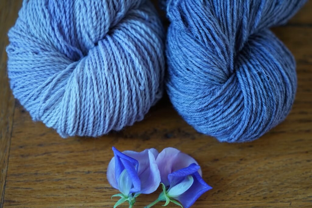 two purple skeins of yarn on a wooden surface next to a blue and purple sweet pea flower