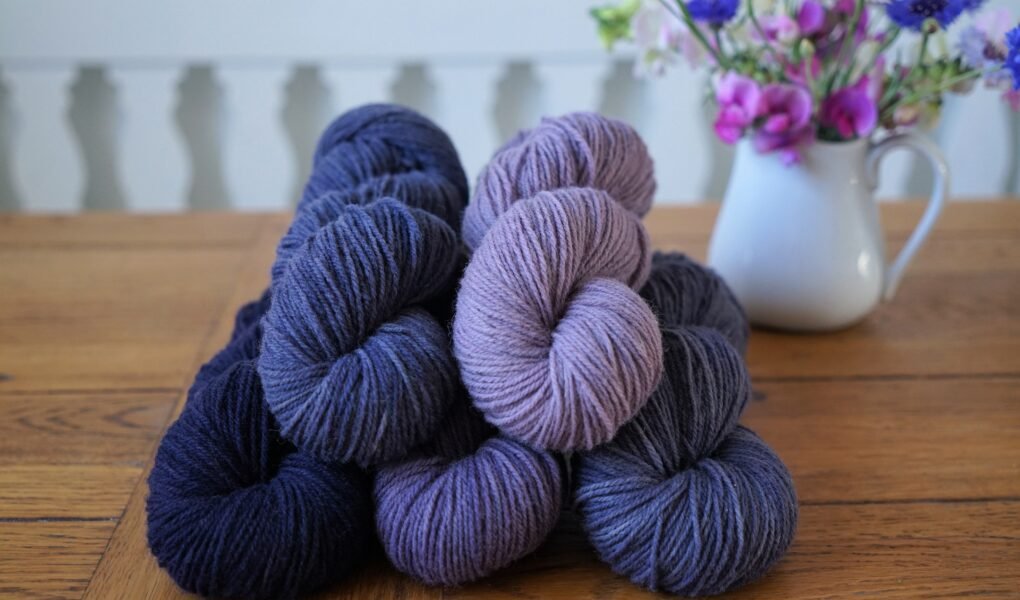 stack of purple and blue skeins on a wooden table and a milk jug filled with flowers
