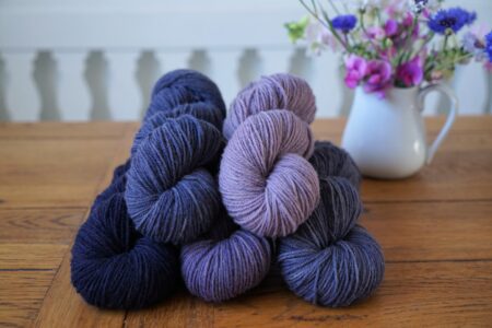 stack of purple and blue skeins on a wooden table and a milk jug filled with flowers