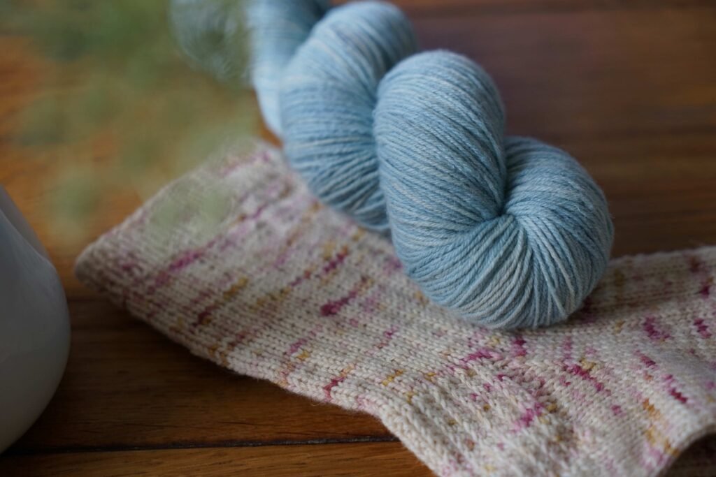 soft blue skein of yarn (Rosemary & Pines Fiber Arts Merino Lino) laying on topf of a speckled socks of the same yarn