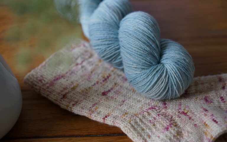 soft blue skein of yarn (Rosemary & Pines Fiber Arts Merino Lino) laying on topf of a speckled socks of the same yarn