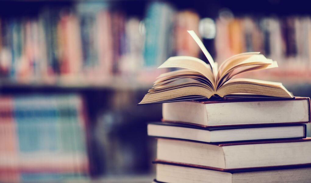 a stack of books in front of a bookshelf, the book at the top is open