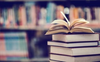 a stack of books in front of a bookshelf, the book at the top is open