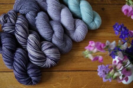 five skeins of yarn in different colorways of purple and blue next to a bouquet of flowers on a wooden surface