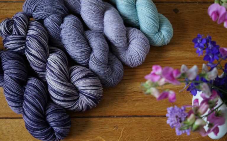 five skeins of yarn in different colorways of purple and blue next to a bouquet of flowers on a wooden surface